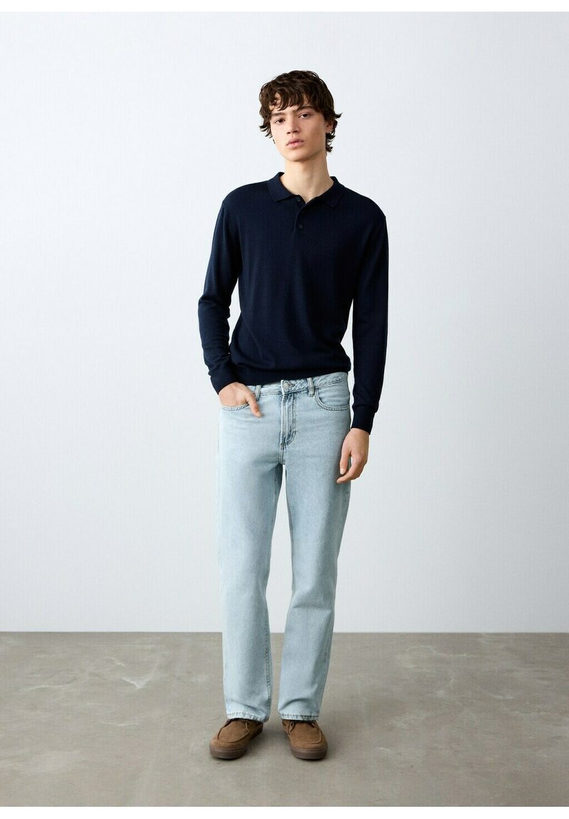 Jeune homme debout portant un polo noir à manches longues, un jean bleu clair et des chaussures marron, devant un mur et un sol gris uni.