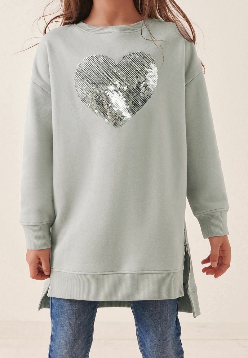 Enfant portant un long sweat gris clair avec un cœur en sequins argentés sur le devant et un jean bleu, debout, les bras détendus sur les côtés.
