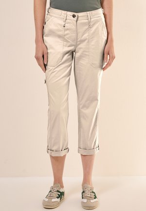 Beige, cropped broeken met zijzakken, opgerolde manchetten en een gladde textuur. Gecombineerd met sneakers met groene accenten.