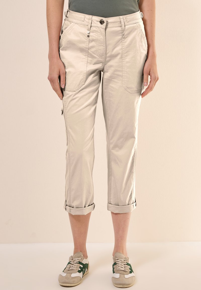 Beige, cropped broeken met zijzakken, opgerolde manchetten en een gladde textuur. Gecombineerd met sneakers met groene accenten.