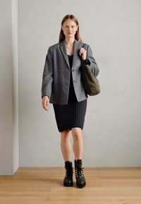 Blazer de mezcla de lana gris, vestido negro de canalé, bolso de hombro verde oliva y botines negros con cordones y suelas gruesas. Suelo de madera, pared gris clara.