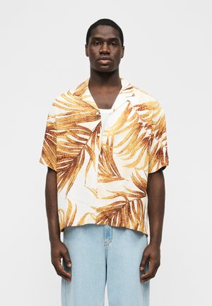 Možakar, oblečen v belo srajco s kratkimi rokavi z rjavim potiskom tropskih listov in svetlo modre kavbojke, stoji pred enobarvnim ozadjem.