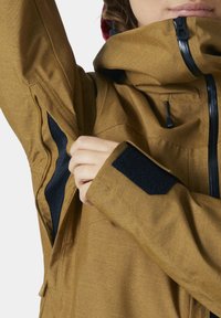 Chaqueta marrón resistente a las inclemencias del tiempo, con un cuello alto, acentos negros y forro de malla. Presenta puños ajustables y bolsillos con cremallera.