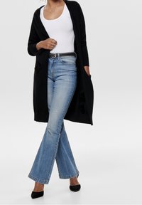 Schwarzer langer Cardigan über einem weißen Tanktop, kombiniert mit hellblauen ausgestellten Jeans und schwarzen spitzen High Heels. Minimalistisches Design mit Taschen.