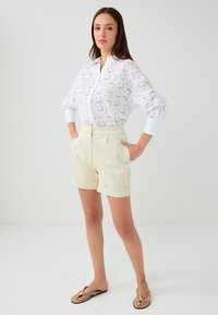Chemise blanche à manches longues avec motif en œillets, associée à un short beige taille haute et des tongs marron. Design simple et ajusté.