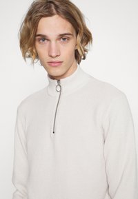 Lys beige strikket genser med høy krage, har en sølvglidelås med en rund trekant, teksturert mønster gjennom hele stoffet.