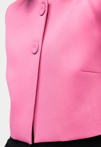 Κοντινό πλάνο ροζ υφασμάτινου σακακιού με δύο καλυμμένα κουμπιά και μια μικρή εγκοπή κοντά στο κάτω άκρο πάνω σε μαύρο παντελόνι.