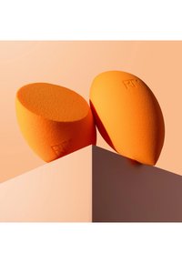 Deux éponges de maquillage orange aux bords arrondis reposant sur une surface géométrique rose clair sur un fond pêche.
