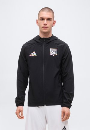 Uomo che indossa una giacca nera con zip con il logo dell'Olympique Lyonnais e il logo Adidas dai colori dell'arcobaleno, pantaloncini bianchi, in piedi davanti a uno sfondo semplice.