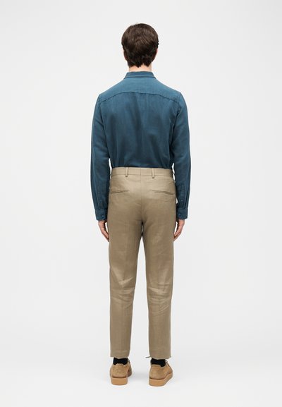 Άνδρας με μπλούζα μακρυμάνικη τιρκουάζ, μπεζ παντελόνι και καφέ παπούτσια, στέκεται γυρισμένος με την πλάτη προς το κάδρο, μπροστά σε απλό ανοιχτό γκρι φόντο.