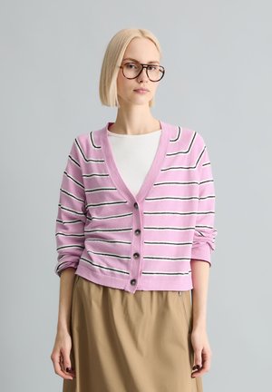 Giovane donna con capelli biondi lisci e occhiali, che indossa un cardigan a righe rosa e bianche con bottoni sopra una maglietta bianca e una gonna beige.