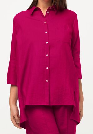 Camisa fucsia abotonada con cuello, mangas largas y un bolsillo en el pecho. Presenta un dobladillo trasero más largo y una textura sutil.