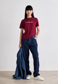 T-shirt en coton bordeaux avec du texte blanc disant "Calvin Klein Jeans", associé à un jean en denim bleu ample et des baskets blanches, tenant une veste en denim.