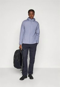 Jasnofioletowa kurtka zapinana na suwak z kapturem, w połączeniu z ciemnymi spodniami i czarnymi butami. Osoba trzyma czarny plecak.