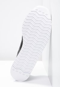 Baskets noires avec une tige texturée, une semelle en caoutchouc blanche présentant un motif ondulé, et un logo embossé sur le côté.