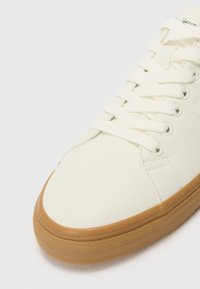 Zapatilla de cuero blanca con punta redondeada, cordones blancos planos y una suela de goma texturada en tono marrón claro. Detalles de costura sutiles en la parte superior.
