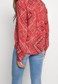 Blouse rouge à motifs géométriques avec accents contrastants, manches longues et coupe ample, associée à un jean bleu clair.