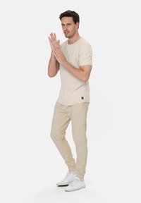 Beige gestreiftes T-Shirt aus weichem Stoff, kombiniert mit hellbeigen Jogginghosen. Weiße Sneaker vervollständigen das Outfit. Das Model befindet sich in einer dynamischen Pose.