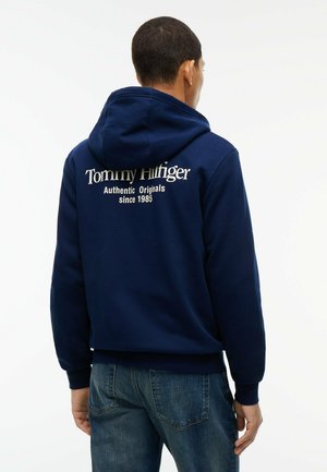 Mann, der mit dem Rücken zur Kamera steht, trägt ein marineblaues Kapuzensweatshirt mit "Tommy Hilfiger Authentic Originals since 1985" auf dem Rücken und blaue Jeans.
