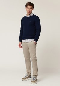 Marineblauwe gebreide trui over een wit gestreept shirt, beige chino's en beige sneakers met blauwe accenten. Model staat tegen een neutrale achtergrond.