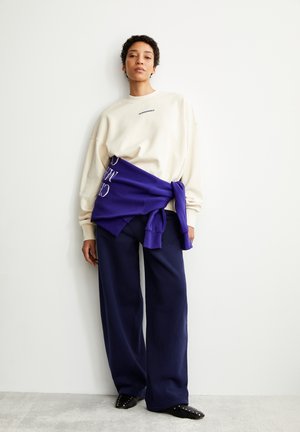 Beige übergroßer Pullover mit lila gebundenem Lagenlook und Text, kombiniert mit navyfarbenen weit geschnittenen Hosen und schwarzen Slippern mit Nieten.