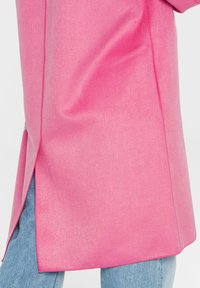 Pink coat with side slit worn over light blue denim jeans, showing fabric texture and seam details.