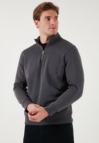 Felpa grigia con collo a zip, maniche lunghe, texture morbida e polsini a costine, abbinata a jeans scuri per un look casual.