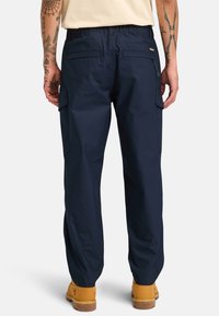 Marina cargo byxor med elastisk midja, sidofickor och avslappnad passform, kombinerade med beige arbetsskor. Förstärkt sömnad synlig.