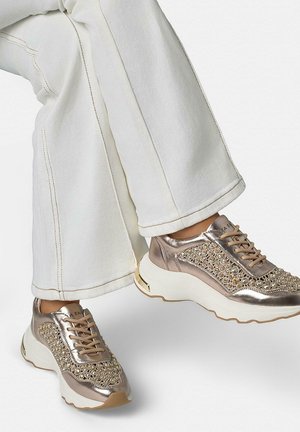 Persona che indossa pantaloni bianchi e sneaker beige metallizzate arricchite con strass e suole spesse bianche.