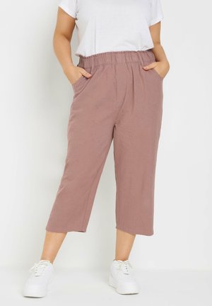 Person iført lilla elastiske bukser med lommer og hvide sneakers, stående mod en hvid baggrund.