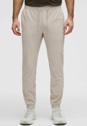 Mand iført beige teksturerede joggebukser, hvide sneakers og en brun skjorte, stående mod en ensfarvet lys baggrund.