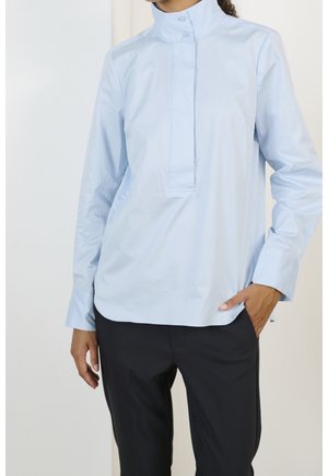 Person iført lyseblå, langærmet top med høj hals og skjult knapstolpe samt sorte bukser, hånd i lommen.