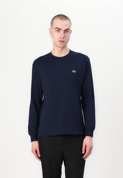T-shirt à manches longues bleu marine en coton, avec un col ras du cou et un petit logo brodé vert sur la poitrine gauche.
