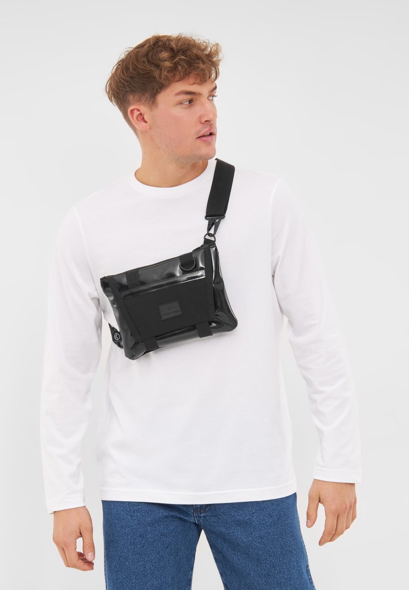 Borsa a tracolla nera, lucida e impermeabile, di forma rettangolare, con tasca anteriore e tracolla regolabile, indossata sopra una camicia bianca a maniche lunghe.