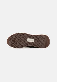 Semelle d'une chaussure en caoutchouc marron avec des rainures diagonales et une étiquette de marque blanche rectangulaire au centre sur fond blanc.