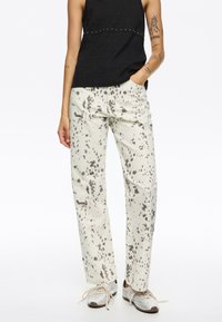 Pantalones blancos con un patrón salpicado en gris, con un corte relajado, bolsillos laterales y un estilo casual, combinados con una camiseta negra ajustada.