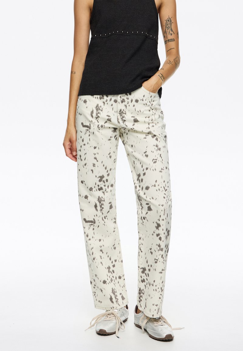 Pantalones blancos con un patrón salpicado en gris, con un corte relajado, bolsillos laterales y un estilo casual, combinados con una camiseta negra ajustada.