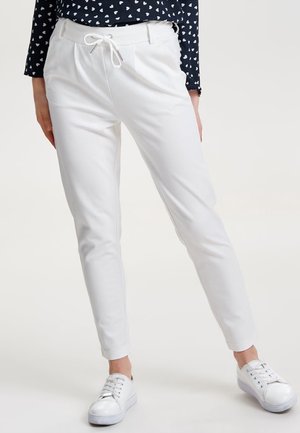 Femme portant un pantalon blanc fuselé avec taille à cordon et des baskets blanches, debout devant un fond uni clair.
