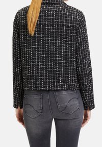 Kobieta z długimi blond włosami, ubrana w czarno-białą kratkę kurtkę i ciemnoszare dżinsy z ozdobnymi tylnymi kieszeniami, pokazana od tyłu.