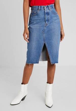Kvinde iført blå denim midi-nederdel med slids foran, rød top og hvide ankelstøvler, stående på lysegråt gulv og baggrund.