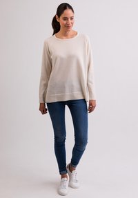 Hellbeige Pullover mit Rundhalsausschnitt, langen Ärmeln und geripptem Saum. Kombiniert mit blauen Skinny Jeans und weißen Sneakern.