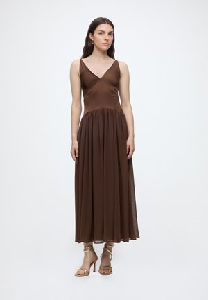 Femme debout portant une robe midi marron sans manches à col en V et des sandales à talons hauts dorées sur un fond clair uni.