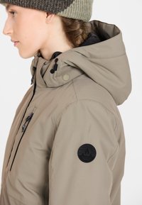 Beige huva jacka med en texturerad tyg, svart dragkedja och ett cirkulärt logomärke på ärmen. Har justerbara dragsko på huvan.