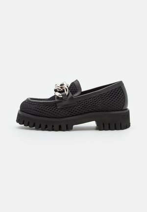 Mocassin noir texturé avec semelle épaisse à crampons et grand détail de chaîne argentée sur le dessus, sur fond blanc uni.