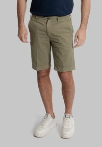 Pantaloni corti in cotone verde oliva con vestibilità dritta, orlo risvoltato e tasche frontali, abbinati a sneakers bianche su uno sfondo neutro.