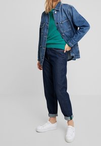 Jeansjacke in blauer Farbe, grüner Pullover, dunkle gerollte Jeans und weiße Sneakers. Merkmale sind zwei Taschen und Knopfverschlüsse.
