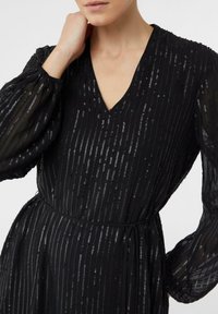 Czarna sukienka z pionowymi paskami, przezroczystymi długimi rękawami, dekoltem w serek i paskiem do wiązania w talii. Teksturowana tkanina z cekinowymi akcentami.