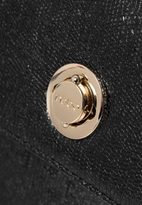 Schwarze handgefertigte Tasche mit einem glatten goldenen runden Verschluss, der den Markennamen trägt und fein detailliert mit einem subtilen geprägten Muster versehen ist.