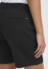 Nærbilde av en person som har på seg svarte shorts med en baklomme og et lite metallmerke med "oxmoc" på høyre side.