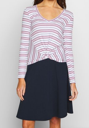 Vrouw draagt een wit shirt met lange mouwen en rode en marineblauwe strepen, geknoopt in de taille, gecombineerd met een marineblauwe rok tot aan de knie.